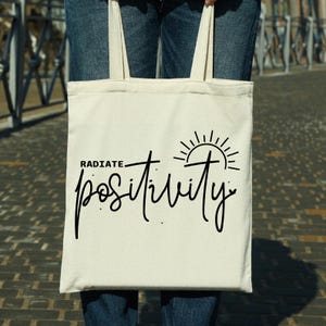 Pu&ograve; includere: Una borsa tote bianca con testo nero che dice "RADIATE POSITIVITY" e un grafico del sole.