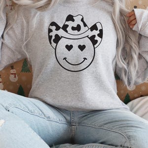 May include: A light gray sweatshirt featuring a smiley face with heart-shaped eyes and a black and white cow print cowboy hat. The design is centered on the front of the long-sleeved top. The sweatshirt is made of a soft material.