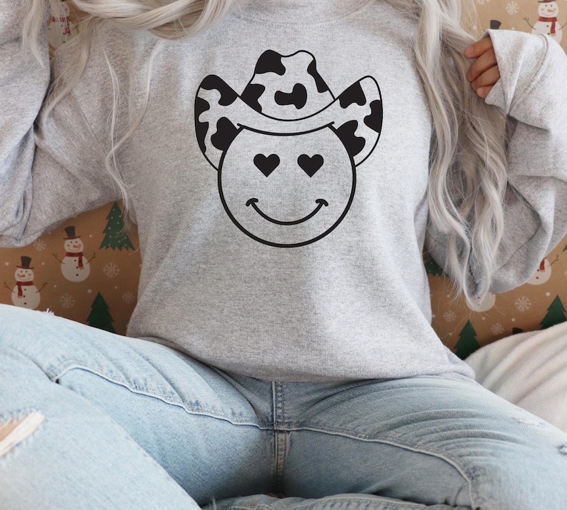 May include: A light gray sweatshirt featuring a smiley face with heart-shaped eyes and a black and white cow print cowboy hat. The design is centered on the front of the long-sleeved top. The sweatshirt is made of a soft material.