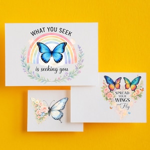 Puede incluir: Tres tarjetas de felicitaci&oacute;n blancas con dise&ntilde;os de mariposas. Una tarjeta presenta una mariposa azul sobre un arco&iacute;ris con el texto "WHAT YOU SEEK is seeking you". Otra tarjeta tiene dos mariposas y el texto "SPREAD YOUR WINGS and Fly."