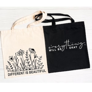 Pu&ograve; includere: Due borse tote, una nera e una beige. La borsa nera ha un testo bianco che dice "tutto andr&agrave; bene". La borsa beige ha un testo nero che dice "diverso &egrave; bello" e un disegno a linea nera di fiori.