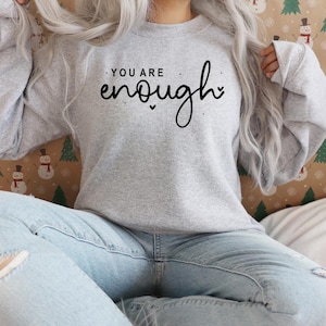 Pu&ograve; includere: Una felpa grigia con il testo "You are enough" in lettere corsive nere.