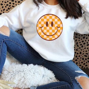 May include: White sweatshirt featuring a large, retro-style smiley face graphic. The smiley face has a checkered pattern in shades of orange and cream, with a brown outline and details. The sweatshirt is a casual, comfortable style.