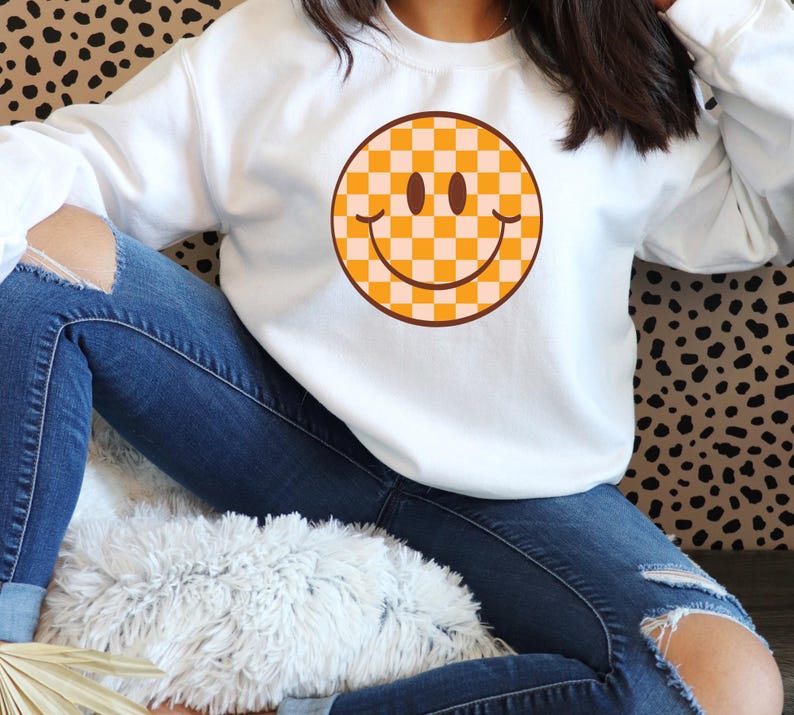 May include: White sweatshirt featuring a large, retro-style smiley face graphic. The smiley face has a checkered pattern in shades of orange and cream, with a brown outline and details. The sweatshirt is a casual, comfortable style.