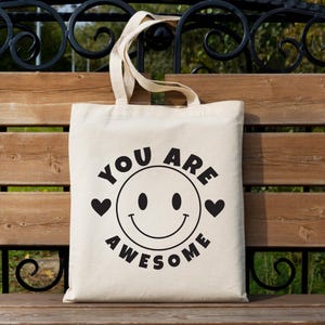 May include: A cream-colored tote bag with black text and graphics. The design features a smiling face with the words "YOU ARE AWESOME" in a circular arrangement. Two black hearts flank the smiley face. The bag has two beige handles.