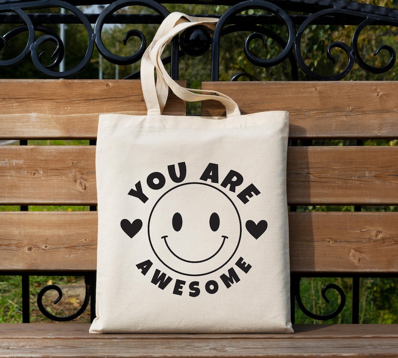 May include: A cream-colored tote bag with black text and graphics. The design features a smiling face with the words "YOU ARE AWESOME" in a circular arrangement. Two black hearts flank the smiley face. The bag has two beige handles.