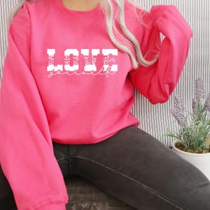 Pu&ograve; includere: Una felpa rosa con le parole "LOVE yourself" in lettere bianche con un motivo leopardato. Le lettere sono in un carattere grassetto e squadrato.