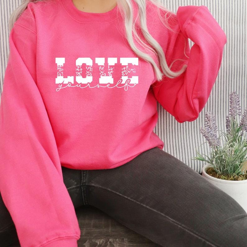 Pu&ograve; includere: Una felpa rosa con le parole "LOVE yourself" in lettere bianche con un motivo leopardato. Le lettere sono in un carattere grassetto e squadrato.