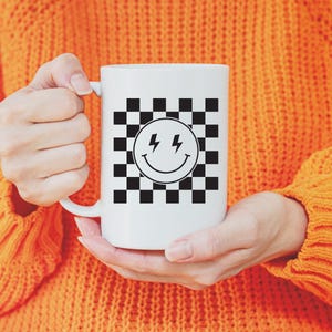 May include: White ceramic mug featuring a black and white checkered pattern with a smiling face and lightning bolt graphic. The mug is held by a person wearing an orange sweater.
