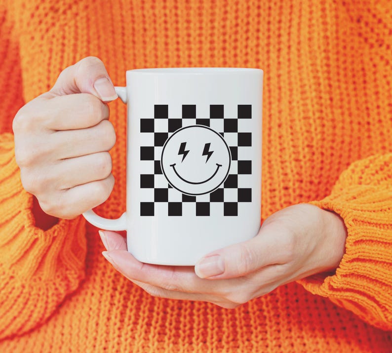 May include: White ceramic mug featuring a black and white checkered pattern with a smiling face and lightning bolt graphic. The mug is held by a person wearing an orange sweater.