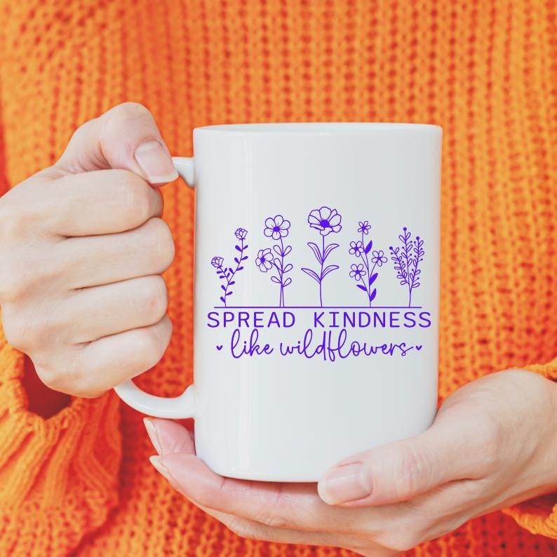 Pu&ograve; includere: Una tazza in ceramica bianca con un design viola di fiori selvatici e il testo "SPREAD KINDNESS - like wildflowers -"