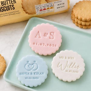 May include: Three scalloped biscuits with embossed text and designs on a light green tray, alongside a package of M&S Food All Butter Biscuits. One pink biscuit reads A & S 01.07.2026, one blue biscuit shows a heart icon with DIVYA & KIRAN 12.10.2026, and one white biscuit reads MR & MRS Willis 01.07.2026.