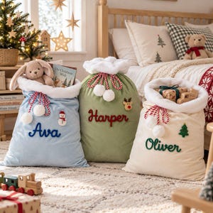 May include: Three soft fabric Christmas gift sacks in light blue, sage green, and cream. Each sack features a white faux fur trim, red and white twisted rope drawstrings with pom-poms, and personalised names embroidered in cursive script.