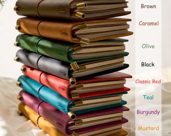 May include: A stack of leather-bound journals in various colors, including brown, caramel, olive, black, classic red, teal, burgundy, and mustard. Each journal has a strap closure and brass accents. The color names are listed on the right side of the image.
