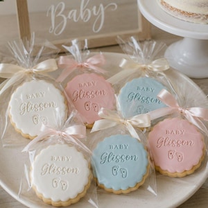 May include: A collection of round, scalloped-edge biscuits individually wrapped in clear plastic bags with light pink ribbons. Each biscuit features the embossed text "BABY Glisson" and a small footprint icon, presented in white, pink, and blue.