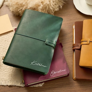 May include: A collection of leather journals in various colors, including green, burgundy, and tan. The green journal is inscribed with the name "Kathleen" in white script. Each journal features a strap closure.