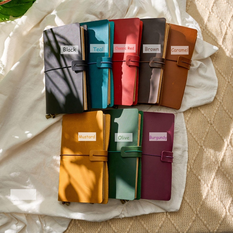 May include: A collection of leather-bound journals in various colors, including black, teal, classic red, brown, caramel, mustard, olive, and burgundy. Each journal is secured with an elastic band and a leather strap closure.