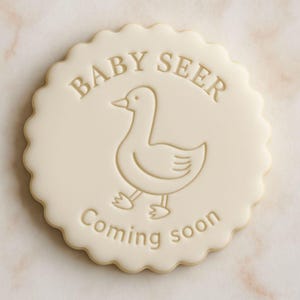 K&ouml;nnte beinhalten: Ein runder, cremefarbener Keks mit gewelltem Rand. Der Keks zeigt ein gepr&auml;gtes Design einer Ente und die Worte "BABY SEER" und "Coming soon". Das Design ist in einem helleren Cremeton gehalten.