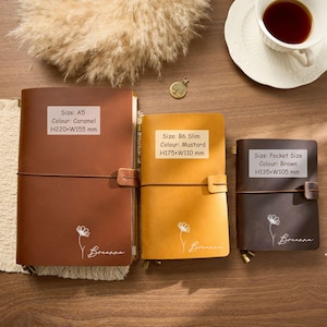 May include: Three leather journals in caramel, mustard, and brown. The caramel journal is A5 size, the mustard is B6 Slim, and the brown is pocket size. Each journal has a floral design and the name "Breanna" printed on the front. The journals are closed with an elastic band.