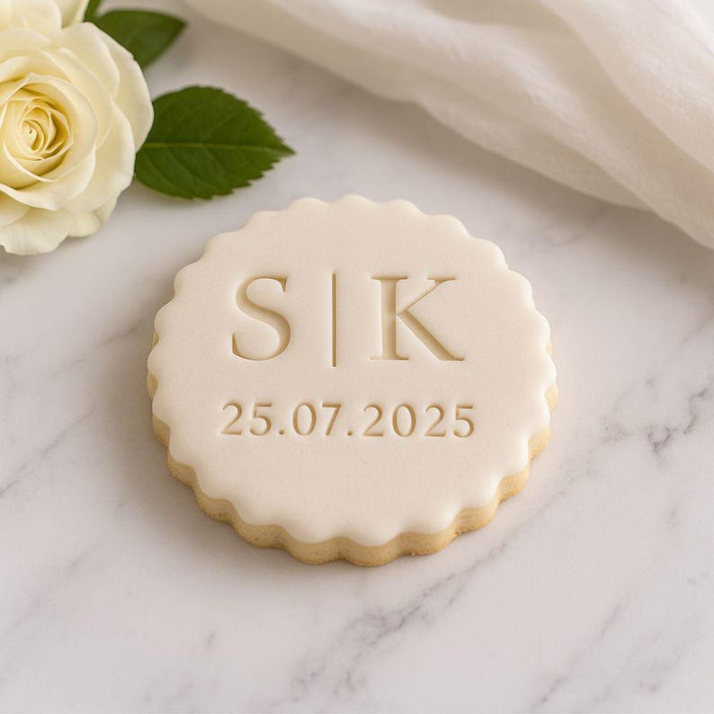 May include: A round, cream-colored cookie with a scalloped edge. The cookie is decorated with the initials "S|K" and the date "25.07.2025". A white rose and green leaf are in the background.