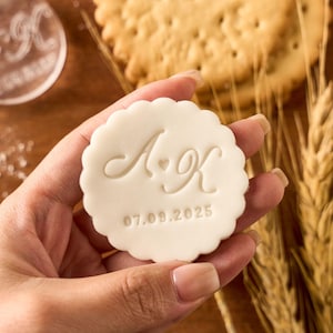 May include: A white, scalloped-edge cookie stamp held in a hand, featuring the initials "A K" with a heart symbol and the date "07.09.2025" embossed. The background includes crackers and wheat stalks.