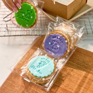 K&ouml;nnte beinhalten: Kekse mit wei&szlig;em Zuckerguss, verpackt in transparenten Zellophanbeuteln. Die Kekse haben einen gewellten Rand und sind mit gr&uuml;ner, lila und blauer Glasur verziert. Die Glasur zeigt den Text "Baby!" und "Oh Baby!" zusammen mit einem Teddyb&auml;ren- und Elefanten-Design.