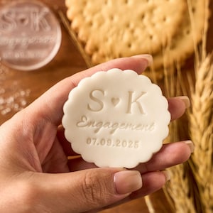 May include: A white, scalloped-edge cookie stamp with the initials "S" and "K" and the word "Engagement" and the date "07.09.2025" engraved on it. The stamp is held in a hand, with a blurred background of crackers and wheat.