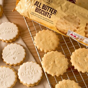 May include: A close-up shot of various biscuits, including round, scalloped-edge butter biscuits and iced cookies. The packaging reads "ALL BUTTER BISCUITS." Some cookies are decorated with the words "Nuna's BIRTHDAY," "BABY Ashton," and "baby OLIVIA."