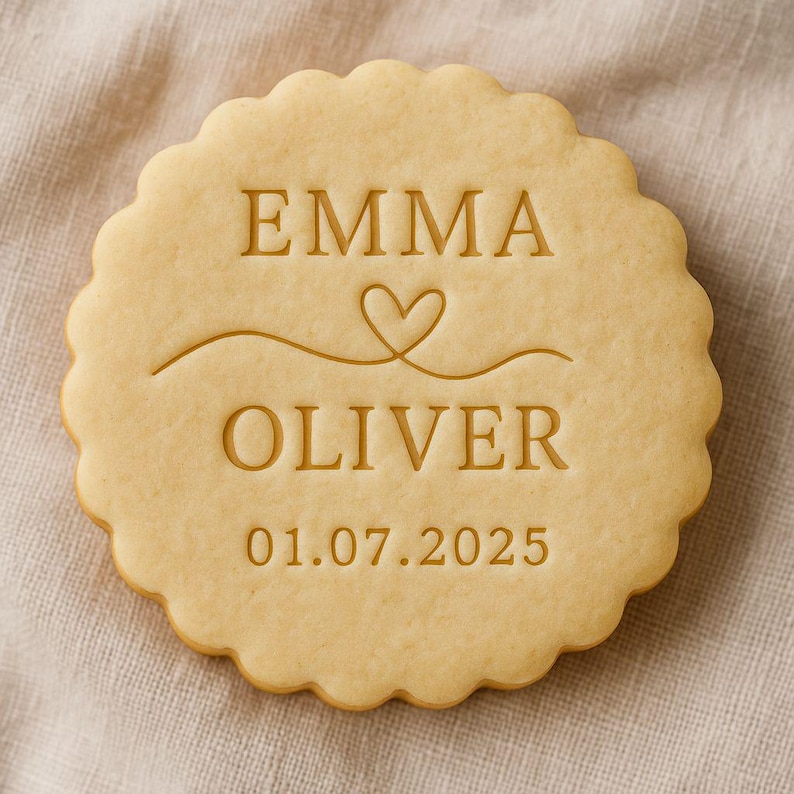 May include: A round, light-brown cookie with a scalloped edge. The cookie is stamped with the names "EMMA" and "OLIVER", a heart symbol, and the date "01.07.2025". The cookie rests on a beige fabric surface.
