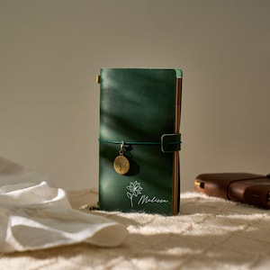 May include: A dark green leather journal with a gold-colored tree charm and a matching elastic closure. The journal is personalized with the name "Melissa" and a floral design. A second brown journal is visible in the background.