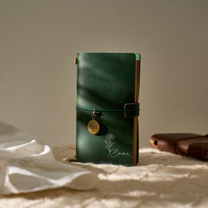 May include: A dark green leather journal with a matching elastic closure and a gold-colored charm. The journal features a floral design and the name "Emma" in white script. A second brown leather journal is visible in the background.