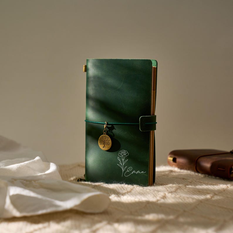 May include: A dark green leather journal with a matching elastic closure and a gold-colored charm. The journal features a floral design and the name "Emma" in white script. A second brown leather journal is visible in the background.
