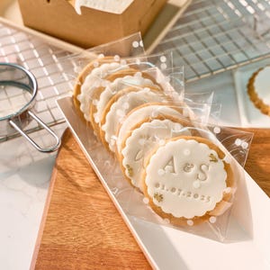 May include: Packaged sugar cookies with white frosting and gold accents. Each cookie is round with a scalloped edge and features the initials "A & S" and the date "01.07.2025". The cookies are in clear cellophane bags with white polka dots.