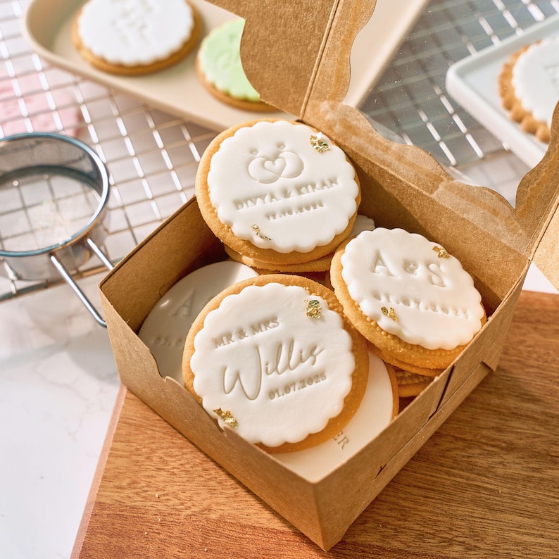 May include: A box of decorated cookies with white icing and gold leaf accents. The cookies have scalloped edges and personalized text, including names and dates. The box is made of brown cardboard and sits on a wooden surface.
