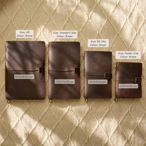 May include: Four brown leather journals of varying sizes are displayed, each with a brass clasp and closure. The sizes are A5, Standard, B6 Slim, and Pocket, with dimensions in millimeters. The journals are arranged on a textured, beige surface.