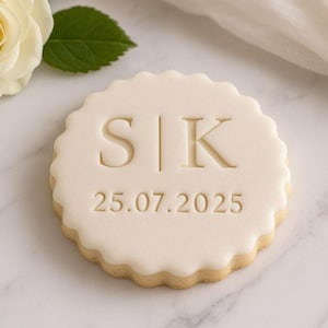 May include: A round, cream-colored cookie with a scalloped edge. The cookie is decorated with the initials "S|K" and the date "25.07.2025" in a simple, elegant font. A white rose and green leaf are in the background.