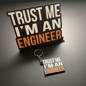 May include: A black and orange keychain with the text "Trust me I'm an engineer" on it. The keychain is attached to a silver key ring.