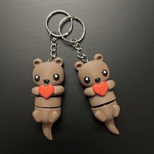 Peut inclure: Deux porte-clés en forme de loutre brune avec des cœurs rouges. Chaque loutre tient un cœur rouge dans ses pattes. Les loutres sont attachées à des porte-clés argentés.