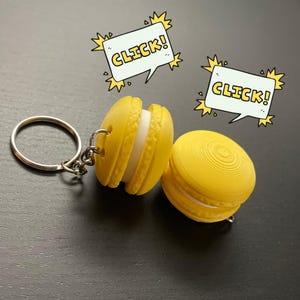 以下が含まれることがあります： 銀色のキーリングが付いた黄色のマカロンキーホルダー。マカロンは白い詰め物が入っており、銀色のチェーンに繋がっています。キーホルダーには「CLICK！」という黄色の文字が書かれたタグが付いています。