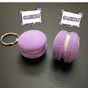 Könnte beinhalten: Zwei lilafarbene Schlüsselanhänger in Macaron-Form mit weißer Füllung. Ein Schlüsselanhänger hat einen silbernen Schlüsselring. Beide Schlüsselanhänger haben eine Sprechblase darüber, auf der "KLICK!" steht.