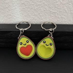 Könnte beinhalten: Zwei avocadoförmige Schlüsselanhänger. Einer zeigt ein rotes Herz, der andere einen herzförmigen Ausschnitt. Beide haben lächelnde Gesichter, rosa Wangen und einen grünen und braunen Rand. Jeder Schlüsselanhänger hat einen silbernen Ring.