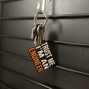 Puede incluir: Un llavero negro y blanco con el texto "Trust me I'm an Engineer" en letras blancas con acentos naranjas. El llavero está unido a un juego de llaves.