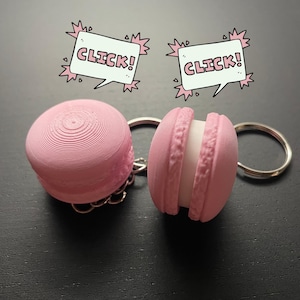 May include: Two pink macaron-shaped keychains with silver key rings. The macarons have a white filling and are attached to the key rings with a silver chain. The text "CLICK!" is written in a speech bubble above each macaron.