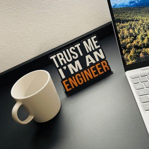 May include: Black metal sign with white text that reads "Trust me I'm an Engineer". The sign is sitting on a black surface next to a white coffee mug.