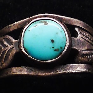 May include: A silver ring with a round, light blue turquoise stone at the center. The ring has an open design with decorative leaf-like patterns on each side of the stone. The ring is set against a black background.