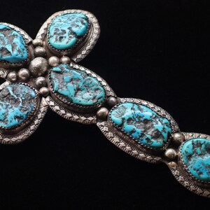 May include: A cross-shaped pendant featuring six oval-shaped turquoise stones with dark matrix patterns. The stones are set in silver bezels with decorative stamped edges and small silver spheres between each stone, attached to a silver chain.