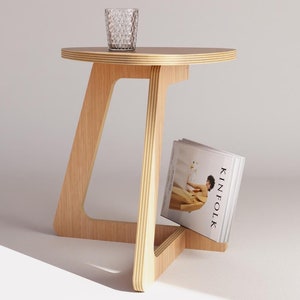 Puede incluir: Una mesa auxiliar de madera clara con una tapa redonda y un estante para revistas debajo. La mesa tiene un diseño único y angulado y está hecha de madera contrachapada.