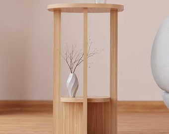 May include: A light wood side table with a round top and a shelf. The table has a vase with dried branches and a glass on top. The table is in front of a light gray sofa.