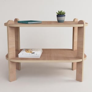 Puede incluir: Una mesa auxiliar de madera clara de dos niveles con bordes redondeados. Una peque&ntilde;a planta en maceta se encuentra en el estante superior, junto con un libro. Una revista est&aacute; en el estante inferior. La mesa es de madera clara.