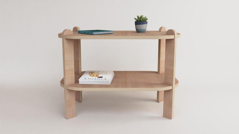 Puede incluir: Una mesa auxiliar de madera clara de dos niveles con bordes redondeados. Una peque&ntilde;a planta en maceta se encuentra en el estante superior, junto con un libro. Una revista est&aacute; en el estante inferior. La mesa es de madera clara.